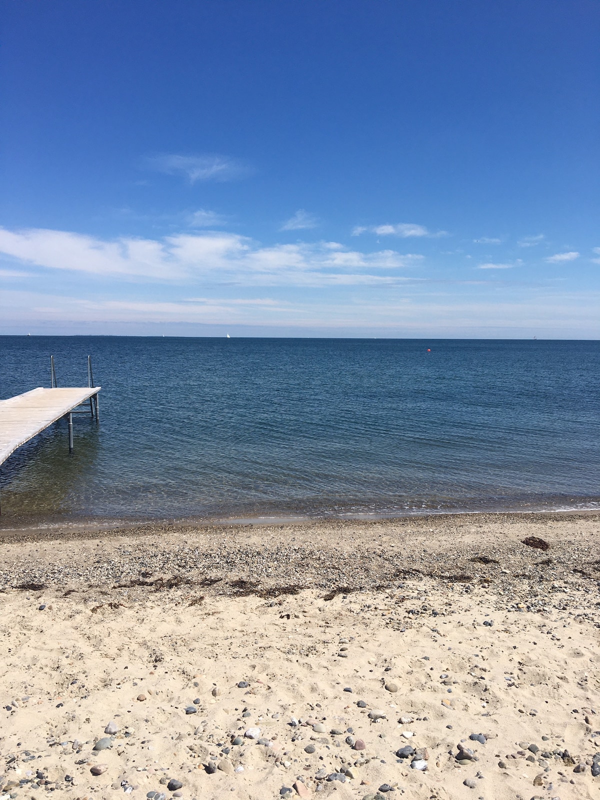 A clear blue sky stretches overhead, reflecting on the calm water of the private beach. A wooden dock extends into the water, surrounded by a sandy shoreline dotted with small stones. The tranquil scenery offers a peaceful view of the distant horizon.