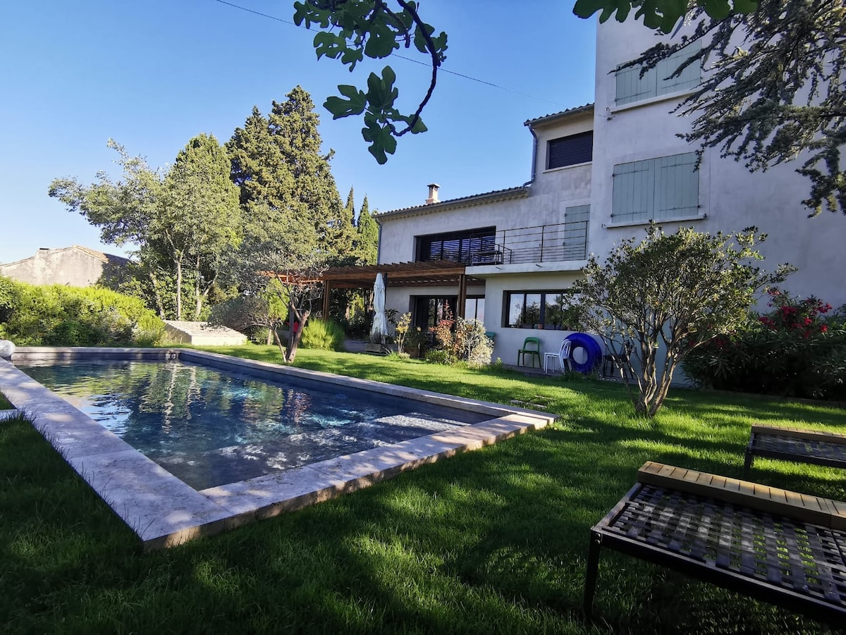A serene garden surrounds a swimming pool, with lush green grass and various plants. The villa features large windows allowing views into the house, while a shaded terrace is visible to one side, complete with outdoor furniture. Trees provide natural shade and character to the setting.