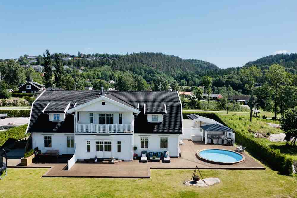 The spacious two-story house features a black-tiled roof and a white exterior. A circular pool is situated in the backyard, surrounded by a landscaped lawn. A terrace with seating is located along one side, providing ample outdoor space for gatherings and relaxation.