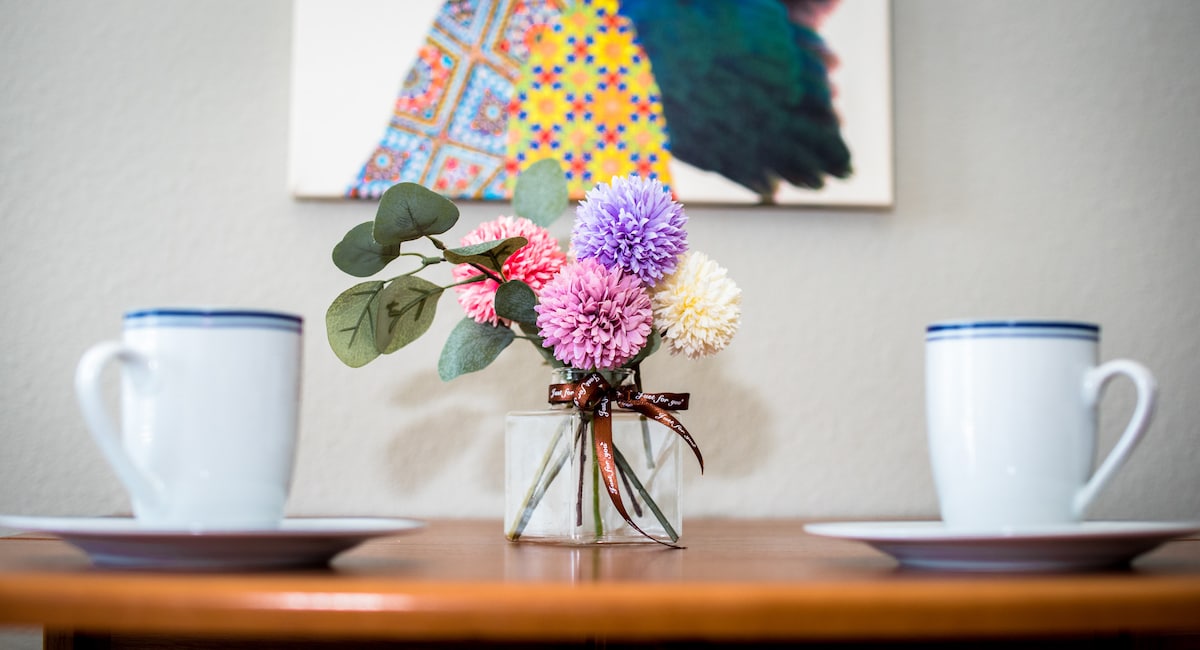 A wooden dining table is set for two, featuring a floral arrangement of colorful blooms in a glass vase. Two white cups with blue rims sit beside coordinating plates, providing a warm and inviting dining setup.