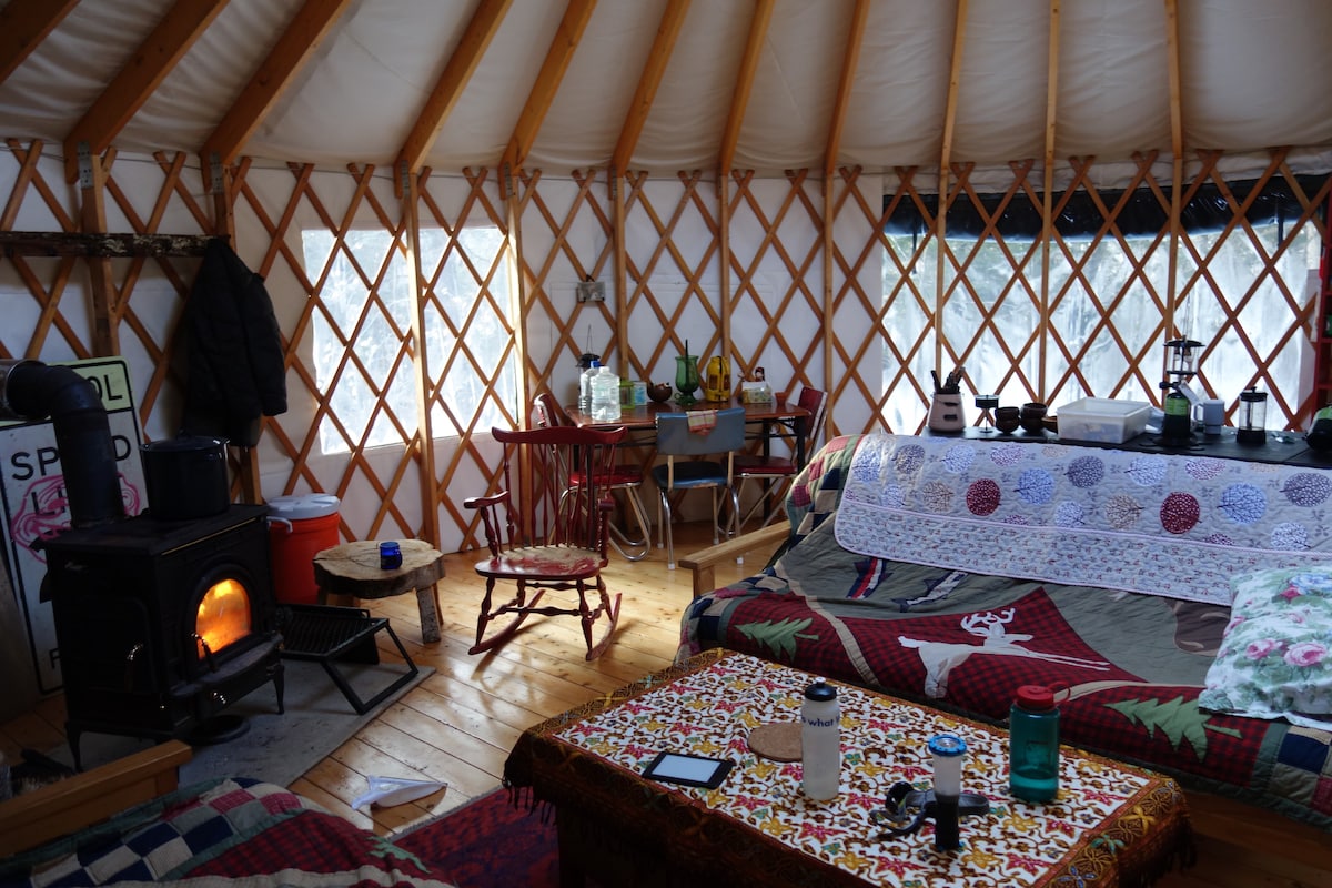 The interior of the yurt features wooden flooring and exposed lattice walls. A wood stove provides warmth, while various kitchen tools and dining accessories are visible on a table. Colorful quilts are draped over the seating areas, creating a cozy environment.