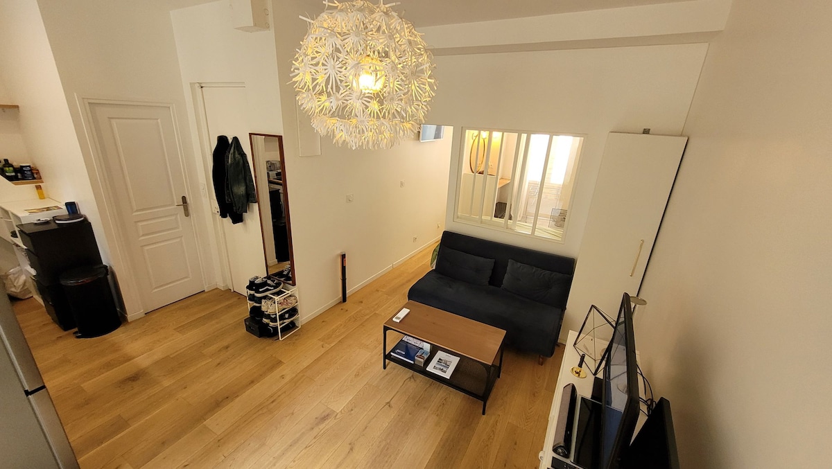 The living area features a modern design, showcasing a dark sofa positioned across from a low wooden coffee table. A TV is visible on a stand, while light filters through large windows, illuminating the wooden flooring. A decorative light fixture hangs from the ceiling.