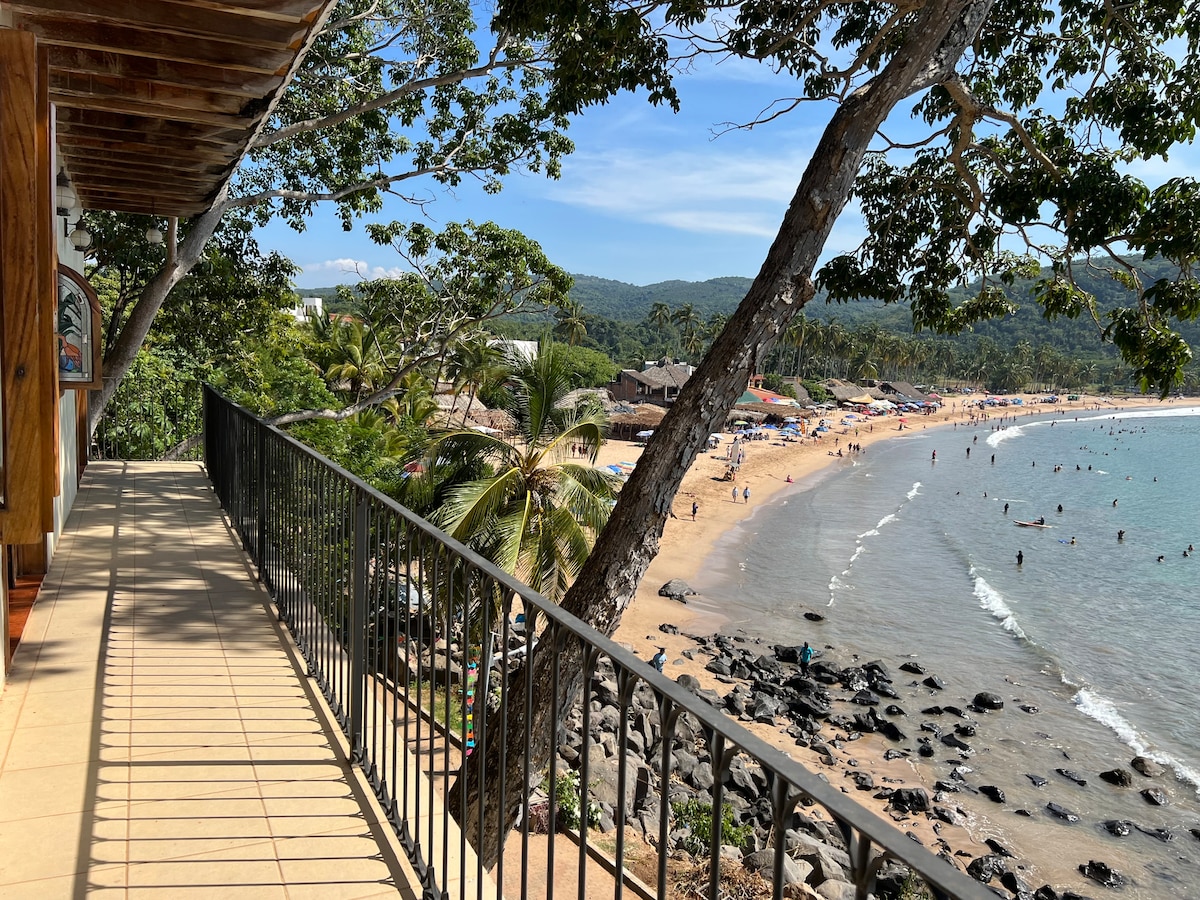 A spacious terrace features a railing overlooking a sandy beach, where people can be seen enjoying the water. Lush green trees provide shade along the walkway, and a vibrant coastline is visible in the distance, framed by gentle waves lapping at the shore.