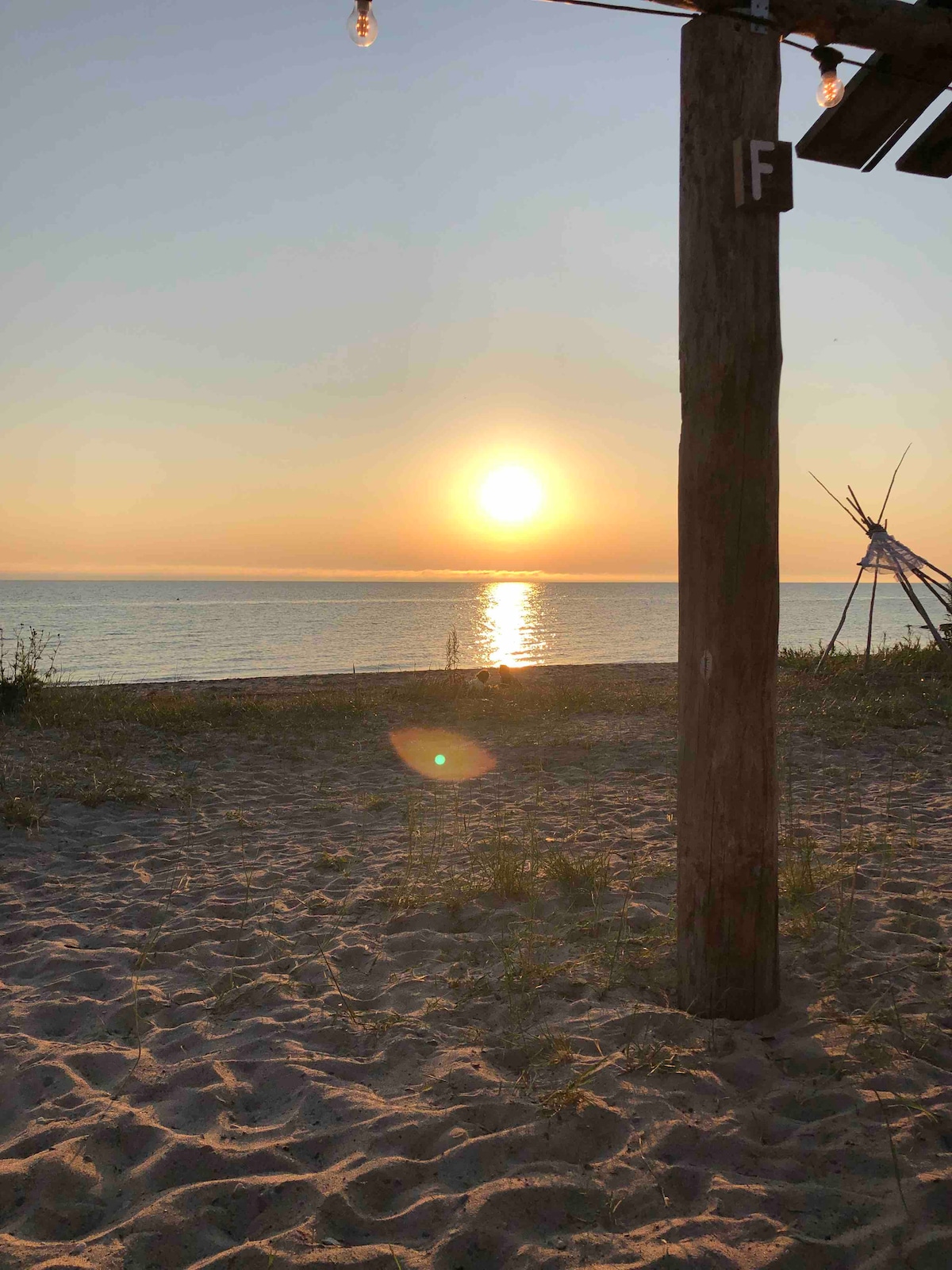 The image captures a serene sunset over the ocean, with the sun reflecting on the water's surface. Soft sand and sparse vegetation frame the foreground. A wooden post stands nearby, contributing to the natural setting. The tranquil scene evokes a sense of calm.