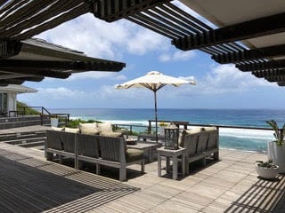 A spacious outdoor lounge area is furnished with comfortable seating, shaded by an umbrella. The wooden decking provides a natural aesthetic, while expansive ocean views create a serene backdrop. Soft colors of the sky and sea complete the tranquil atmosphere.