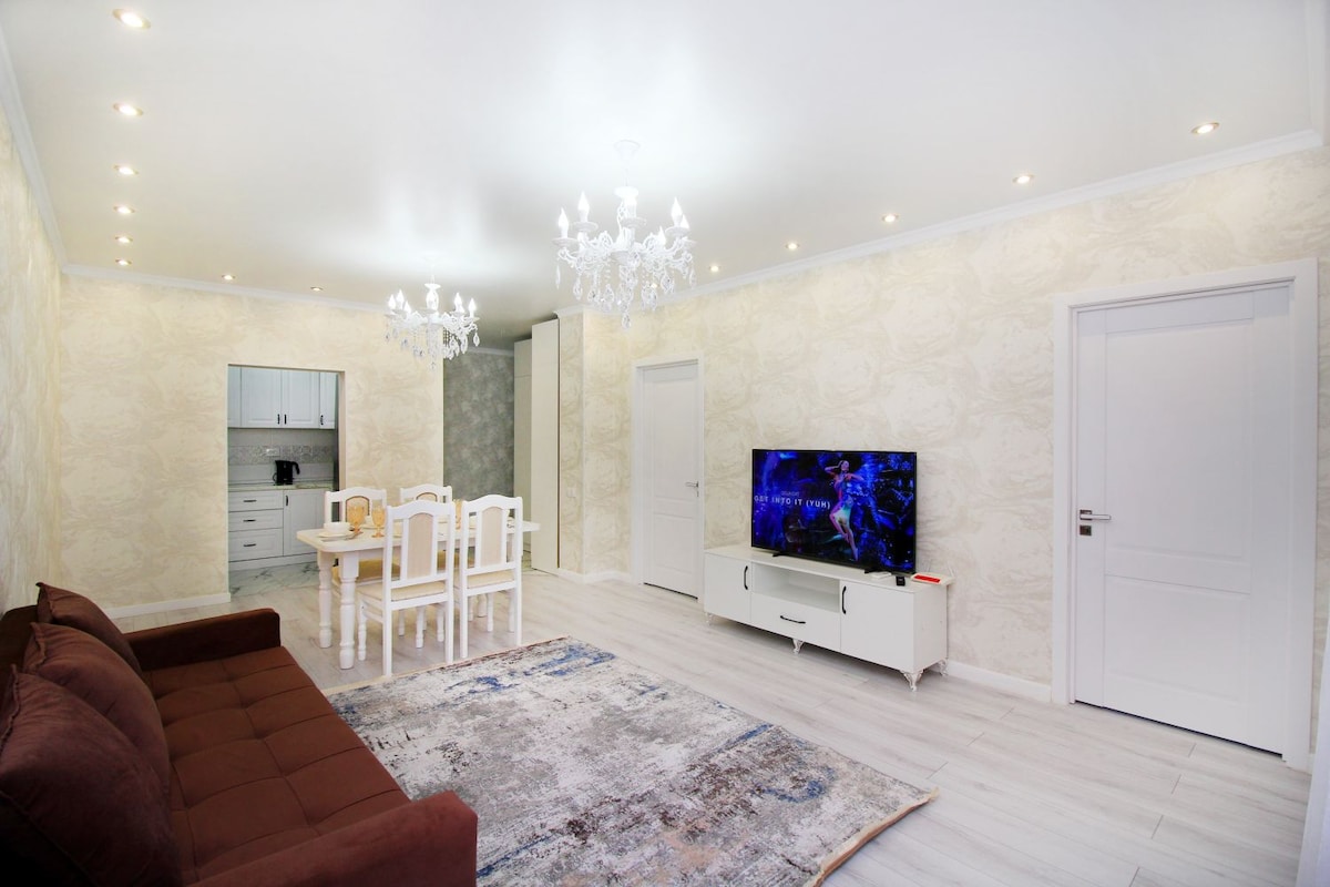 A spacious living area showcases a dark sofa and a white dining table set for four. A large TV resides on a white media console against a light wall. Bright ceiling lights illuminate the room, which features a decorative chandelier and light-colored flooring.