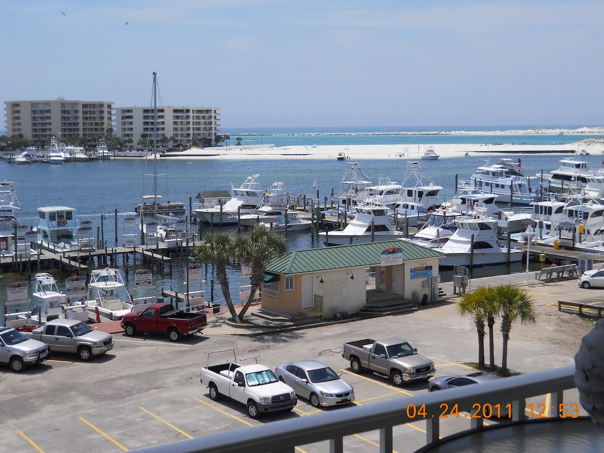 A view of a marina filled with boats docked along a wooden pier. Several vehicles are parked in the lot in the foreground, with palm trees providing a touch of greenery. The distant shoreline and an expansive sky create a serene backdrop to the scene.