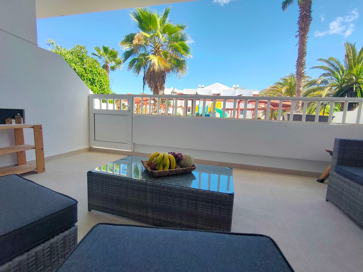 A shaded terrace is furnished with woven lounge seating, accompanied by a low coffee table holding a bowl of fresh fruit. Lush palm trees and clear blue skies are visible in the background, contributing to a relaxing outdoor space.