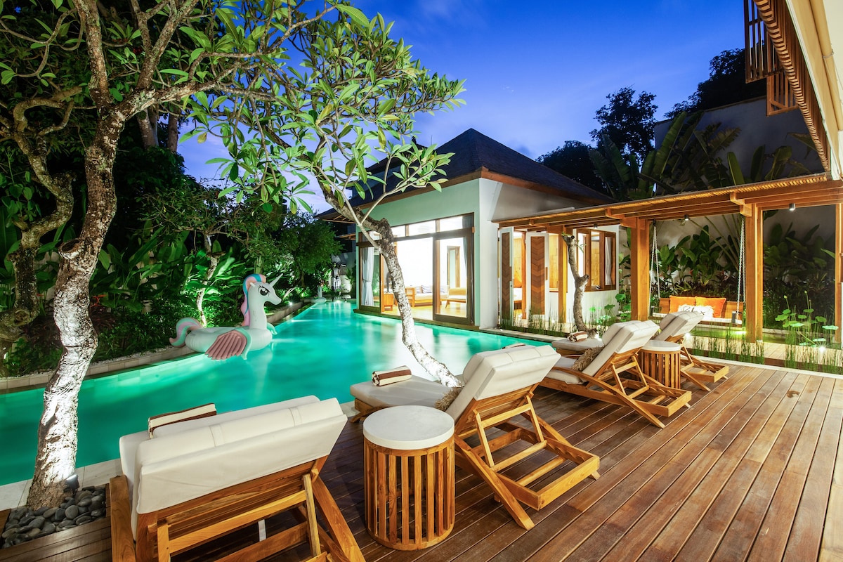 An inviting outdoor space features a private infinity pool surrounded by lush tropical greenery. Sun loungers are arranged on a wooden deck near the pool, with a playful unicorn float adding a whimsical touch. Soft lighting enhances the tranquil atmosphere for evening relaxation.