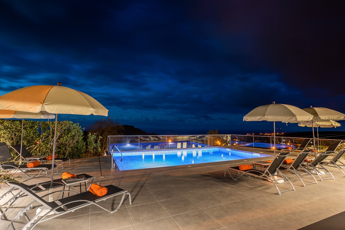 An outdoor swimming pool is illuminated under a night sky, surrounded by several lounge chairs and umbrellas. The pool area is bordered by a glass railing, offering views of the distant hills and city lights.
