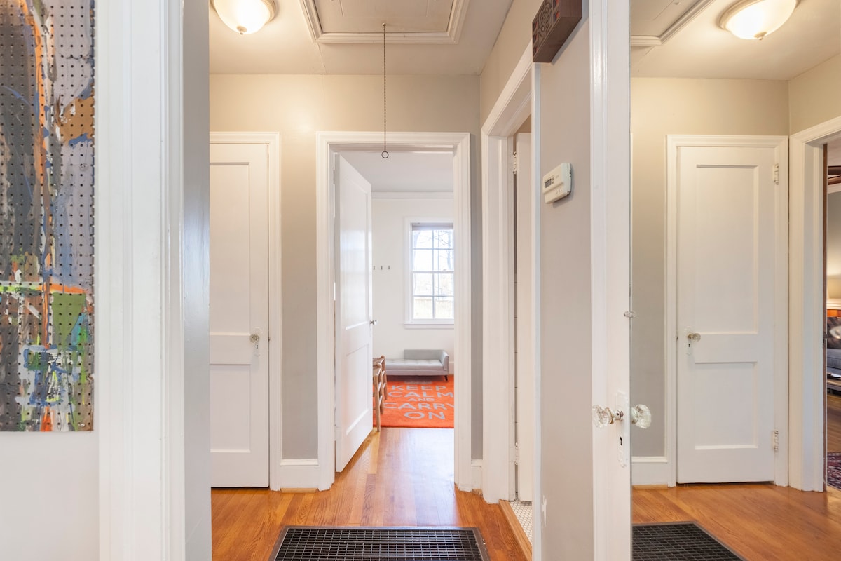 A view of the hallway showcases wooden floors and light gray walls, adorned by framed art. Multiple doors lead to various rooms, while a large window allows natural light to fill the space. Two mats are positioned at the entrances for added convenience.