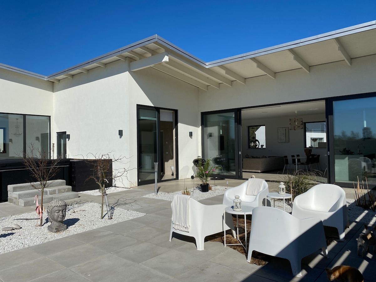 An outdoor seating area features modern white chairs arranged on a stone patio, surrounded by minimalist landscaping with gravel and small plants. Large glass doors offer a view into the interior, inviting natural light into the space. A clear blue sky is visible overhead.