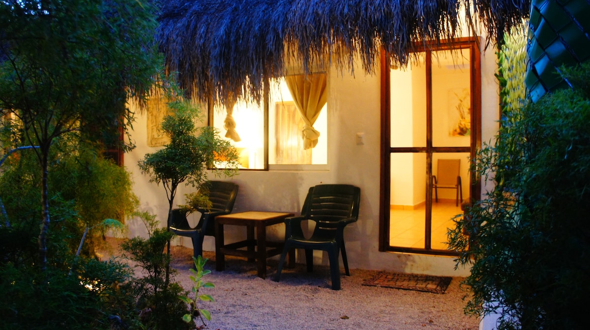 A tranquil outdoor seating area is framed by lush greenery. Two dark chairs and a wooden table are positioned in gravel, with warm light spilling from the windows, creating an inviting atmosphere. The natural thatched roof adds a rustic character to the space.