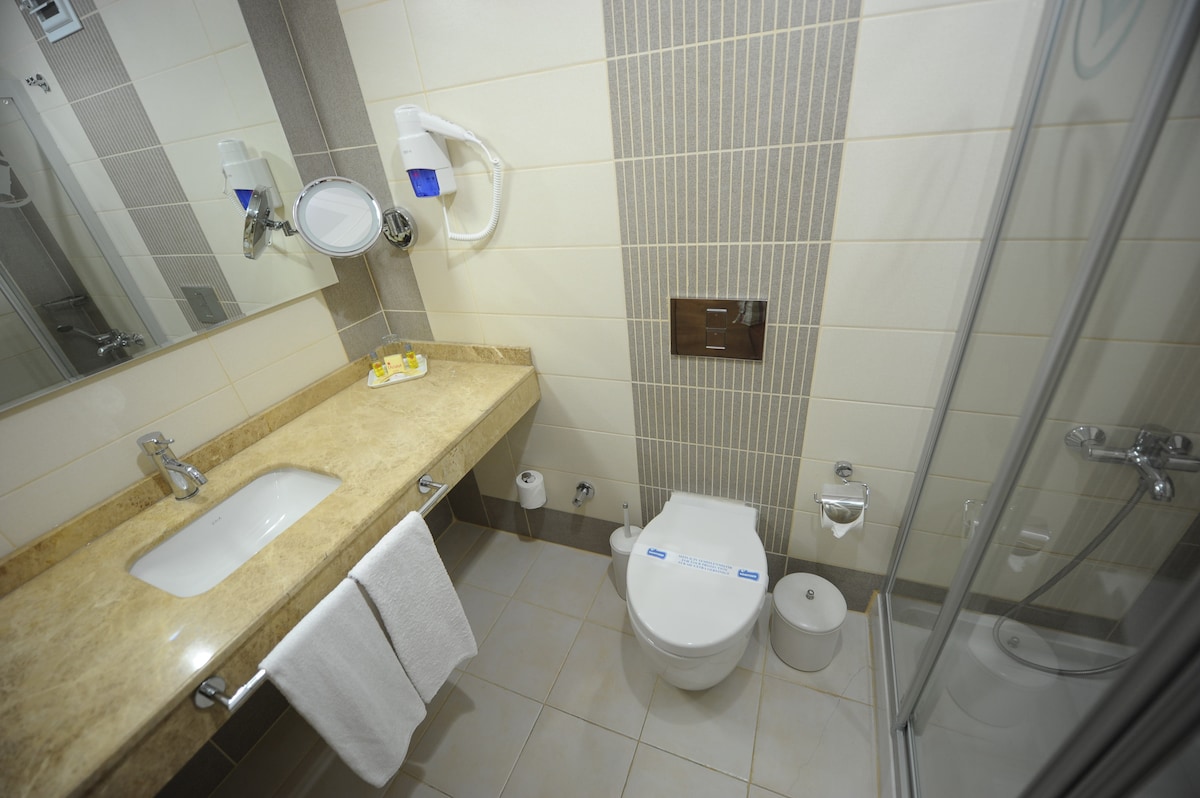 A modern bathroom is equipped with a neutral-toned countertop and a spacious mirror. A white toilet is located beside a glass-enclosed shower. Complementary toiletries are neatly arranged on the counter, with white towels hanging on a bar near the sink.