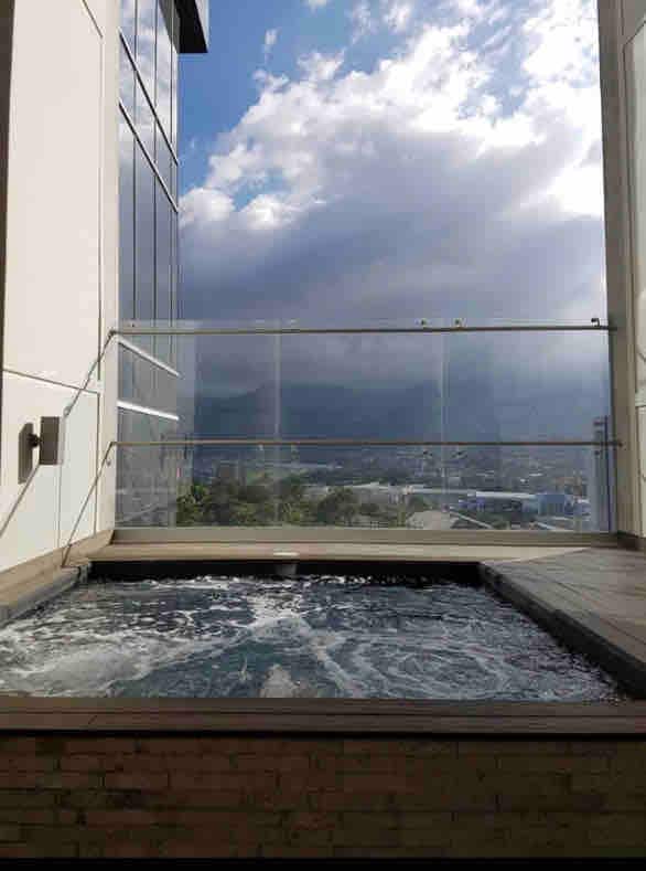 A private hot tub is positioned on a balcony, providing panoramic views of the surrounding city and mountains. The water is rippled, reflecting the sky above, while a glass railing ensures clear sightlines. The deck is made of natural wood, enhancing the outdoor space.