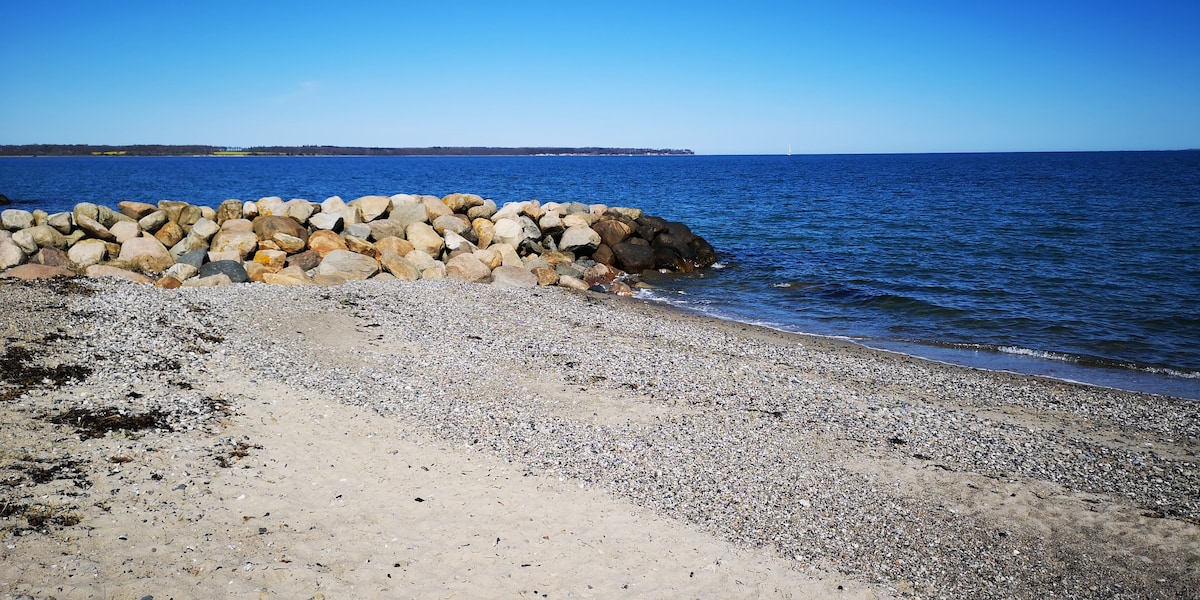 A sandy beach stretches into the distance, framed by a rocky shoreline on one side. The calm blue water reflects the clear sky, with a gentle wave pattern visible. Small pebbles scatter across the shore, enhancing the natural setting.