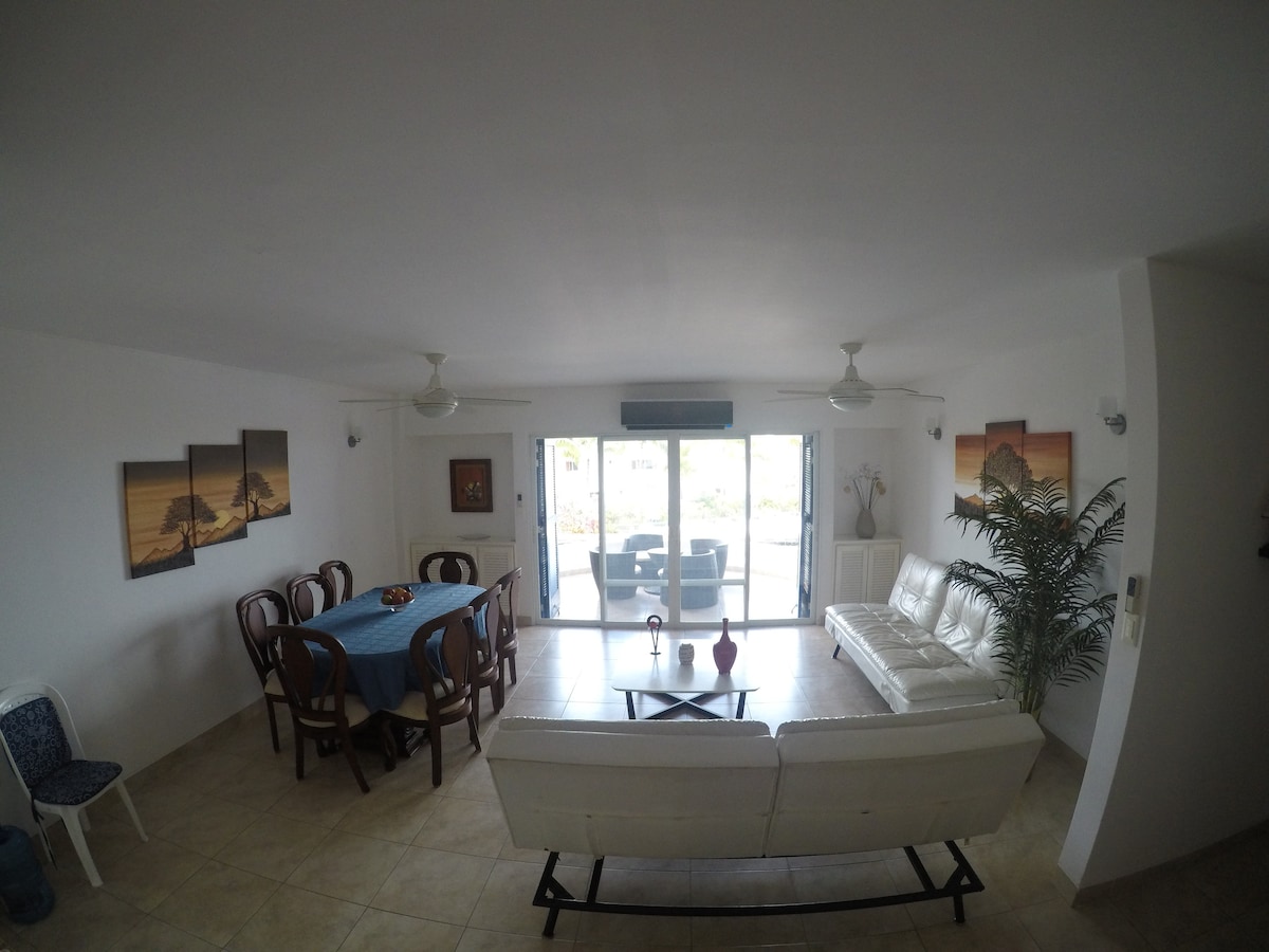 A spacious living area is presented, featuring a white leather sofa and a wooden dining table set with seating for six. Large glass doors open to a terrace, allowing natural light to fill the room. Tropical wall art and a potted plant add subtle decoration.