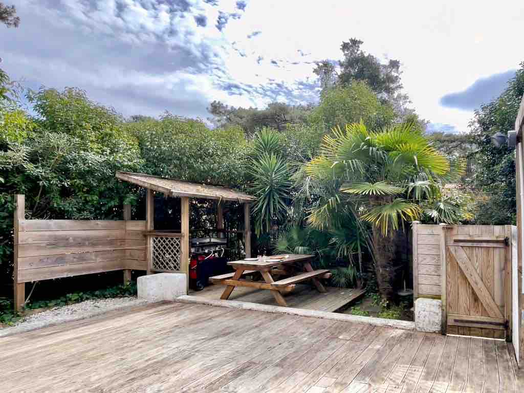A wooden deck is surrounded by lush greenery, featuring a shaded picnic area with a large table. Tropical plants add a touch of nature's beauty, while a cozy covered space is visible in the background, providing sheltered seating.