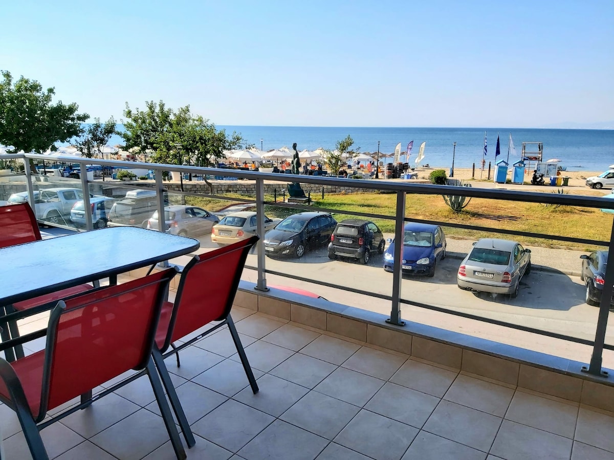 A spacious balcony is furnished with a table and red chairs, providing a view of the sandy beach and the sea. Cars are seen parked along the promenade, complemented by umbrellas and sun loungers in the background.