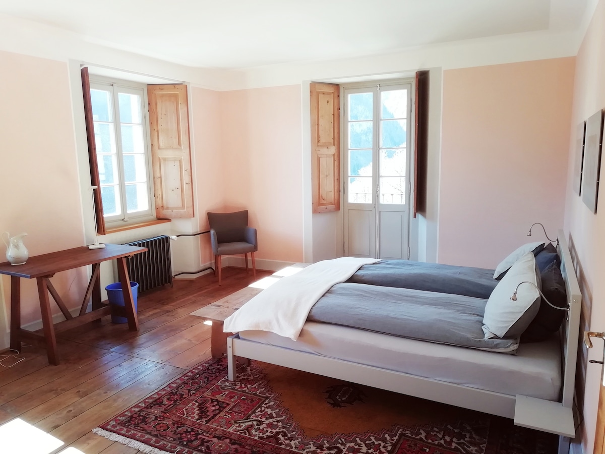 A spacious bedroom features a large bed with gray linens, accompanied by a wooden table and a comfortable chair. Natural light enters through multiple windows with wooden shutters, highlighting the pale pink walls and wooden flooring.