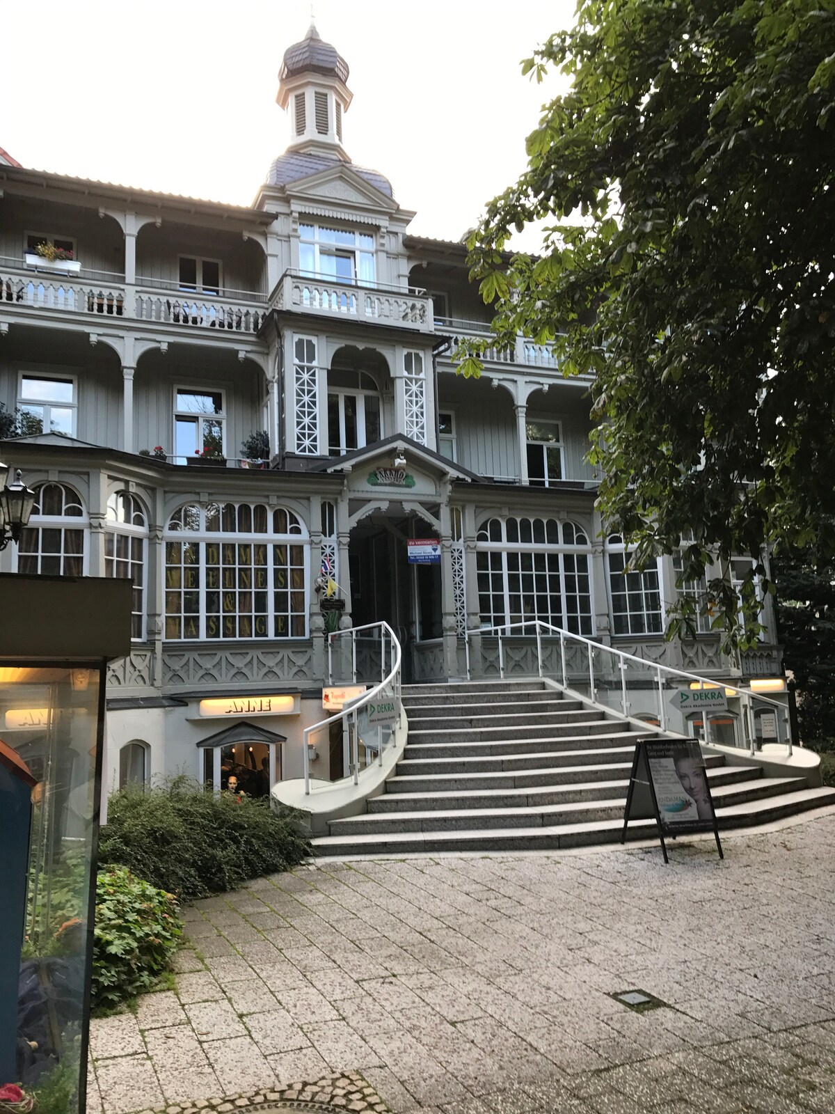 A grand building features a decorative architectural style with multiple balconies and a prominent turret. A wide staircase leads to the entrance, surrounded by lush greenery and walkways. The setting reflects a welcoming atmosphere in a charming neighborhood.