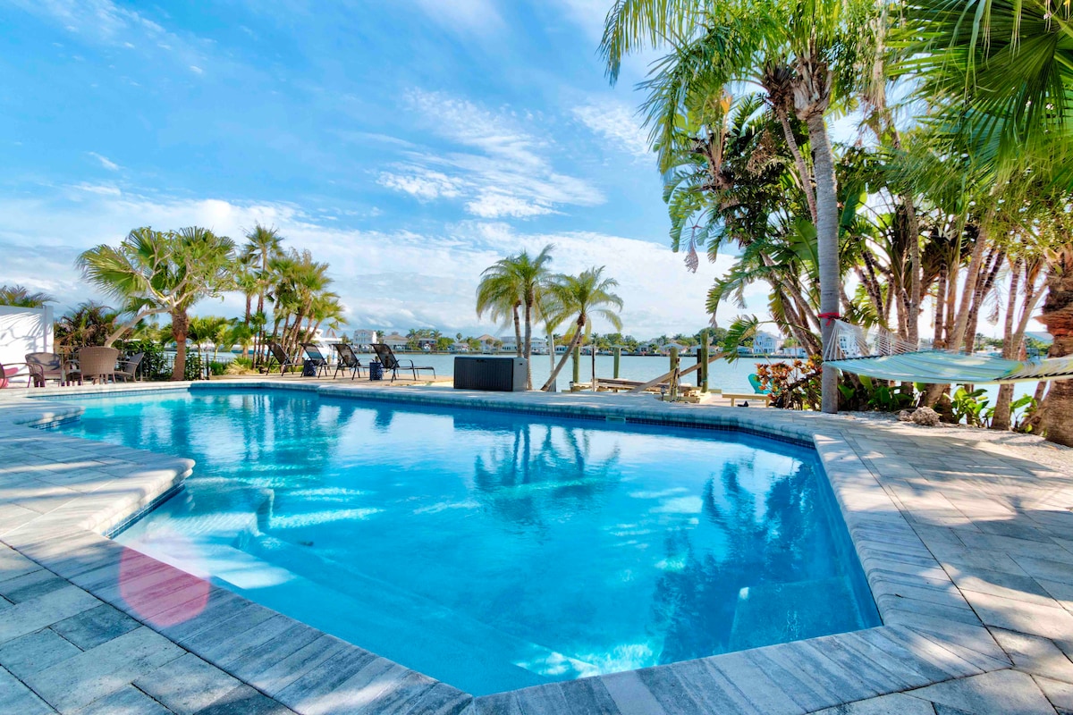 The outdoor pool, surrounded by palm trees, reflects the clear blue sky. Comfortable loungers are placed nearby, offering ample space for relaxation. The glistening water leads to views of the waterfront, enhancing the serene atmosphere of the outdoor environment.
