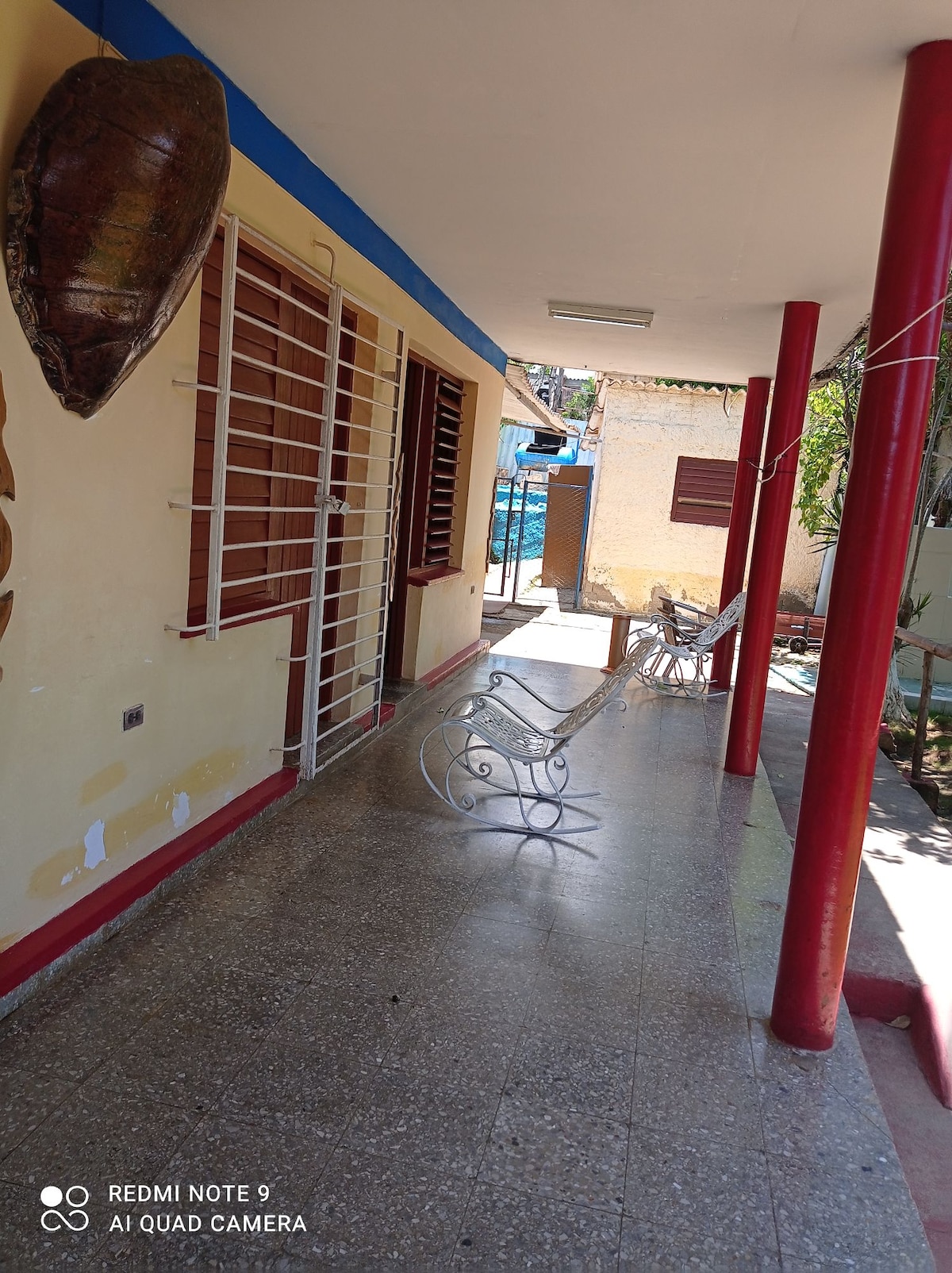 A covered porch area is displayed with red columns and a smooth floor. A white rocking chair is positioned near the entrance, alongside light-colored walls and windows with bars, leading to a vibrant outdoor space beyond.