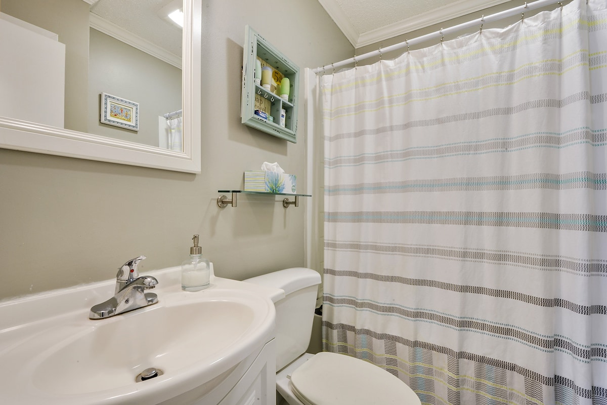 A light-colored bathroom is shown, featuring a white sink and toilet. A shower area with a striped curtain is visible, and a small shelf displays decorative items. Light is provided by overhead fixtures, creating a bright and airy atmosphere.