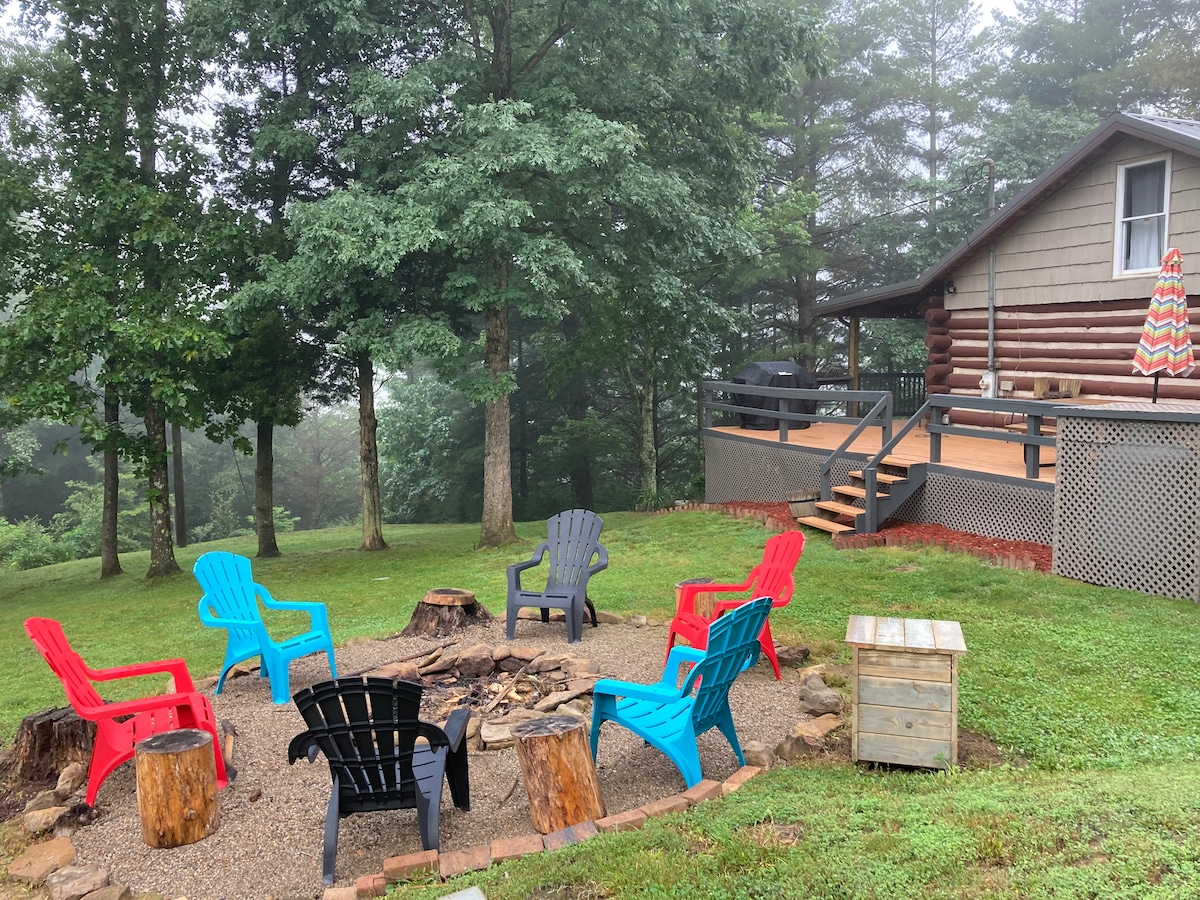 A fire pit area is surrounded by colorful chairs in red, blue, and black, arranged in a circular formation on gravel. A wooden stump serves as a table, with a cabin and deck visible in the background, framed by lush trees. Fog adds a serene atmosphere.