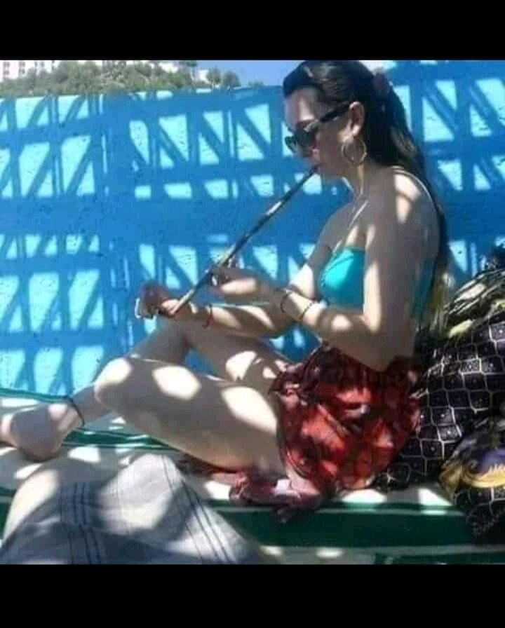 A person is seated on a green bench, playing a flute. The background features a blue lattice structure, casting intricate shadows. A patterned bag sits nearby, while a blanket drapes across the bench. Light filters through the lattice, enhancing the scene.
