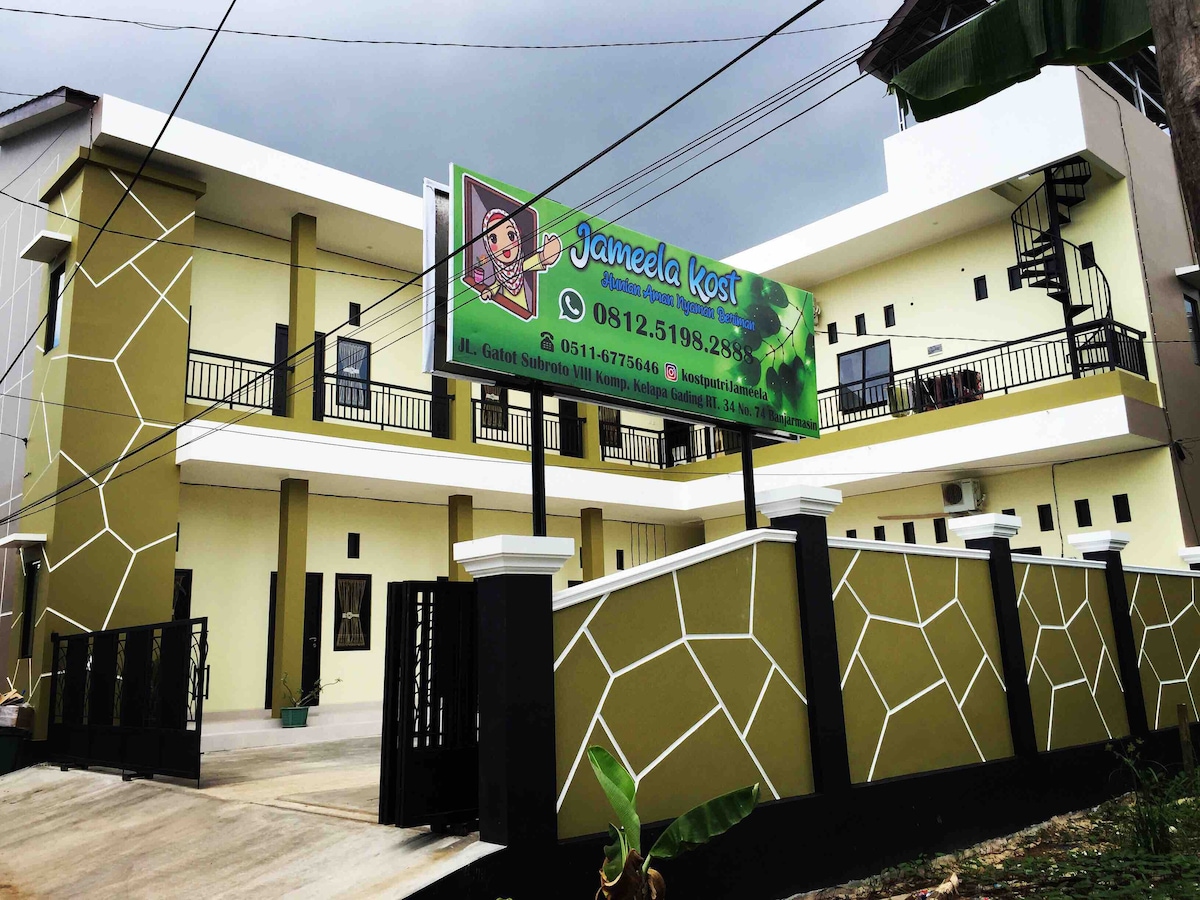 The exterior view of a newly constructed building features a modern design with a prominent sign displaying the name 'Jameela Kost'. A well-defined entrance area is visible, framed by geometric patterns and offering access to the upper floors via exterior staircases. The surrounding area is landscaped.