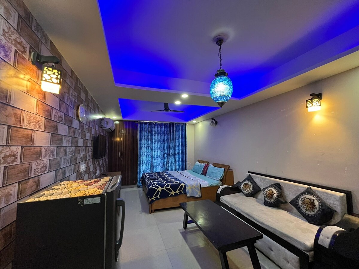 A cozy room features a spacious bed and a sofa adorned with decorative pillows. Soft blue lighting sets a calming tone, highlighting the patterned wall. A small table stands in front of the sofa, while a refrigerator sits against one wall.