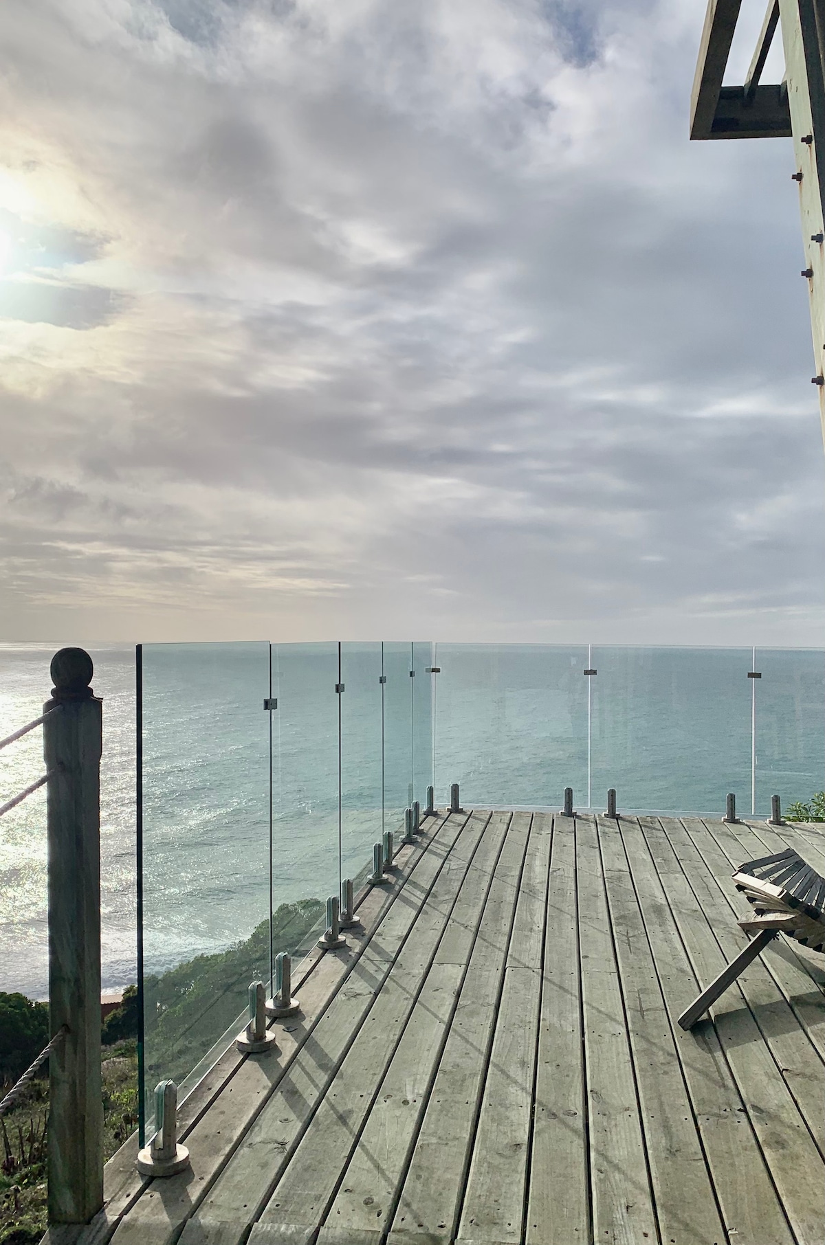 A spacious terrace is presented with a clear glass railing, offering unobstructed views of the ocean waves. The wooden deck features a single wooden chair positioned thoughtfully, inviting relaxation while enjoying the coastal scenery. The sky reflects soft shades of blue and gray, enhancing the serene ambiance.
