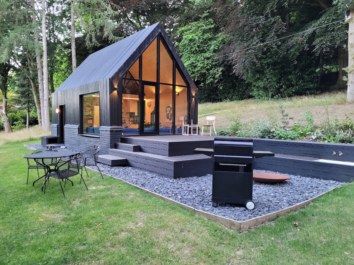 A modern tiny home with a distinctive angular roof is set amidst greenery. The exterior features large glass windows reflecting warm light from the interior. Outdoor dining furniture and a gas BBQ are positioned on a level deck, surrounded by a gravel pathway and landscaped yard.