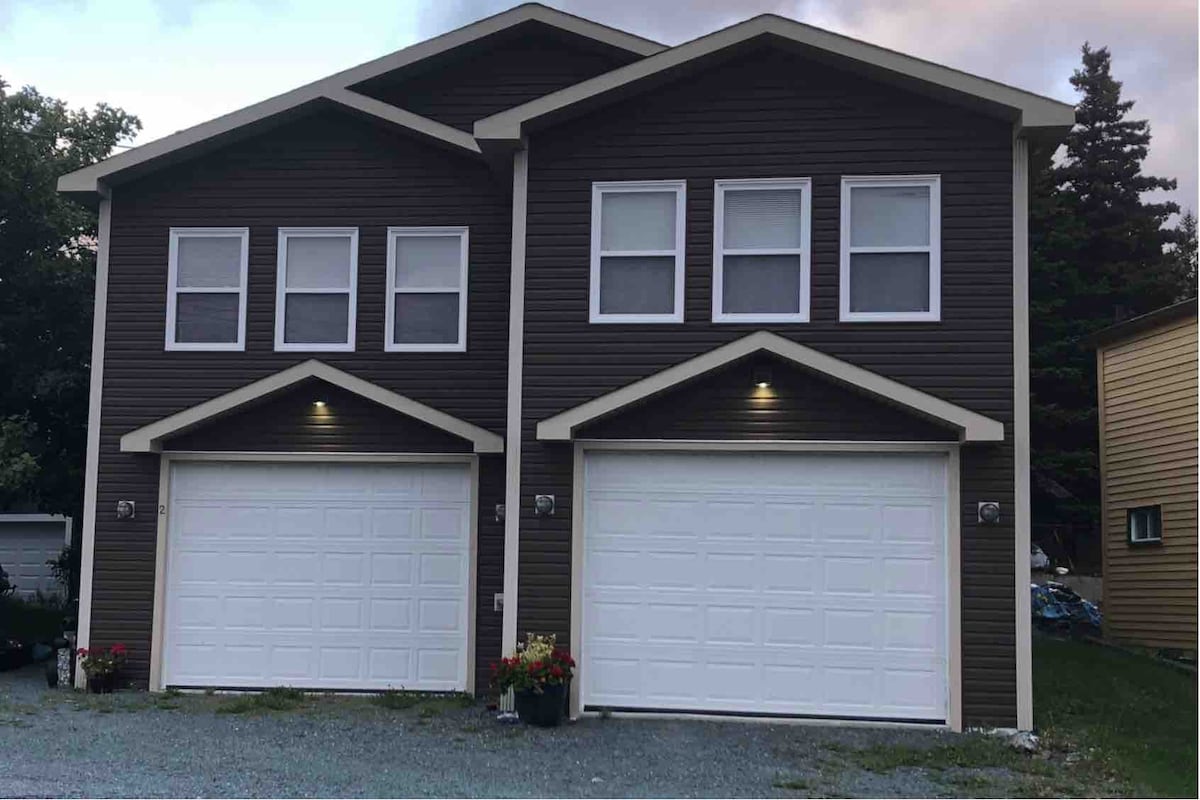 A modern two-story dwelling is shown, featuring a symmetrical facade with large windows and a welcoming design. Dual white garage doors with outdoor lighting provide convenience, while flower planters add a hint of color against the dark exterior.