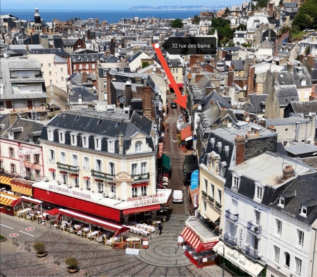 Aerial view of a lively street, showcasing cafés and shops along the cobblestone path. The red awnings of an outdoor dining area and the rooftops of buildings create a charming urban landscape, with a glimpse of the sea visible in the distance.