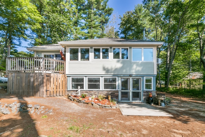 The exterior of the house is visible, featuring a light grey façade with multiple windows allowing natural light inside. A wooden deck is seen on the left, surrounded by trees. A patio area with stone accents is also present, offering potential for outdoor gatherings.