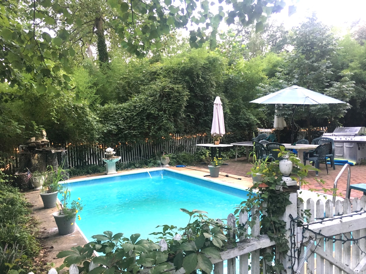 A serene backyard features a heated in-ground pool, surrounded by lush greenery and mature trees. Comfortable seating is positioned near the water, with an umbrella providing shade. A patio area includes a grill, while potted plants add a touch of nature.