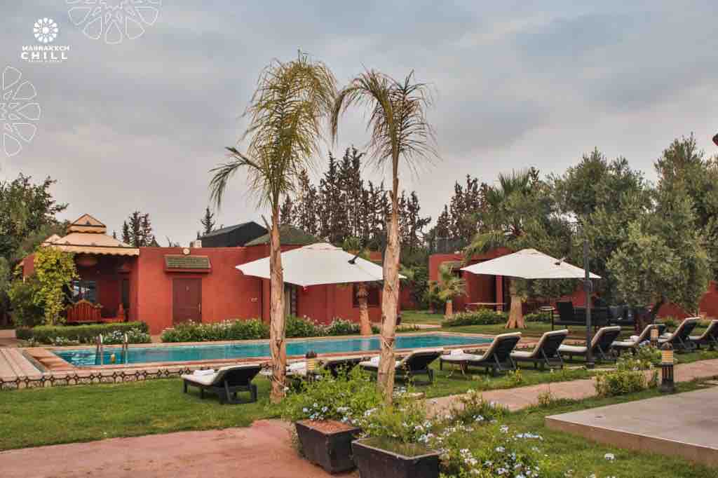 A serene outdoor area showcases a swimming pool surrounded by lounge chairs under white umbrellas. Lush greenery, including palm trees and shrubs, frames the scene, while several adjoining structures provide a warm, inviting backdrop.
