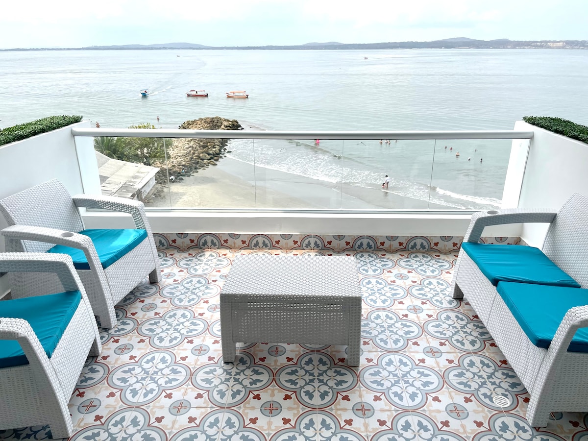 A balcony area features two white wicker chairs with blue cushions and a matching coffee table. The vibrant tiled floor adds a decorative touch. The view showcases the ocean and beach below, with boats in the distance and people enjoying the shore.