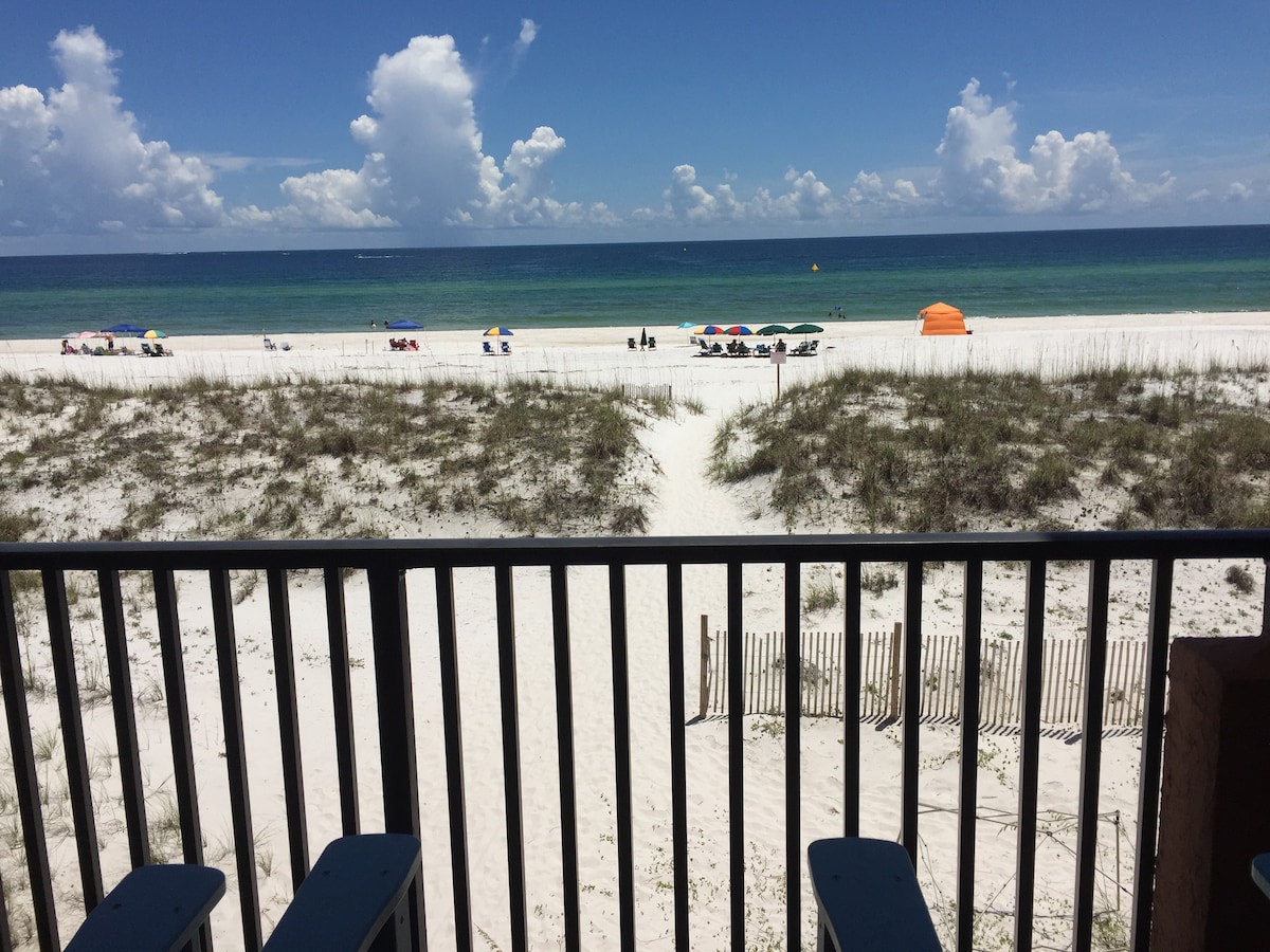 A view from the balcony showcases sandy shores leading to the Gulf of Mexico, where the turquoise waters meet a clear blue sky. Sunbathers are positioned on beach towels, while umbrellas dot the shoreline. Grassy dunes are visible in the foreground, with a wooden fence marking the beach access.