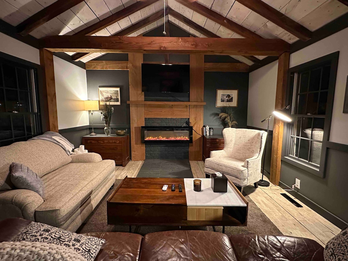 A warm living area is highlighted by exposed wooden beams and a vaulted ceiling. A comfortable sofa and an armchair are positioned around a coffee table. A modern fireplace serves as the focal point, with a television mounted above it, while soft lighting enhances the cozy atmosphere.
