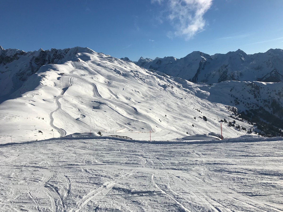 A vast snow-covered landscape stretches across the image, with gentle slopes and trails visible. Majestic mountains rise in the background under a clear blue sky, while the foreground displays a smooth, undisturbed expanse of snow.