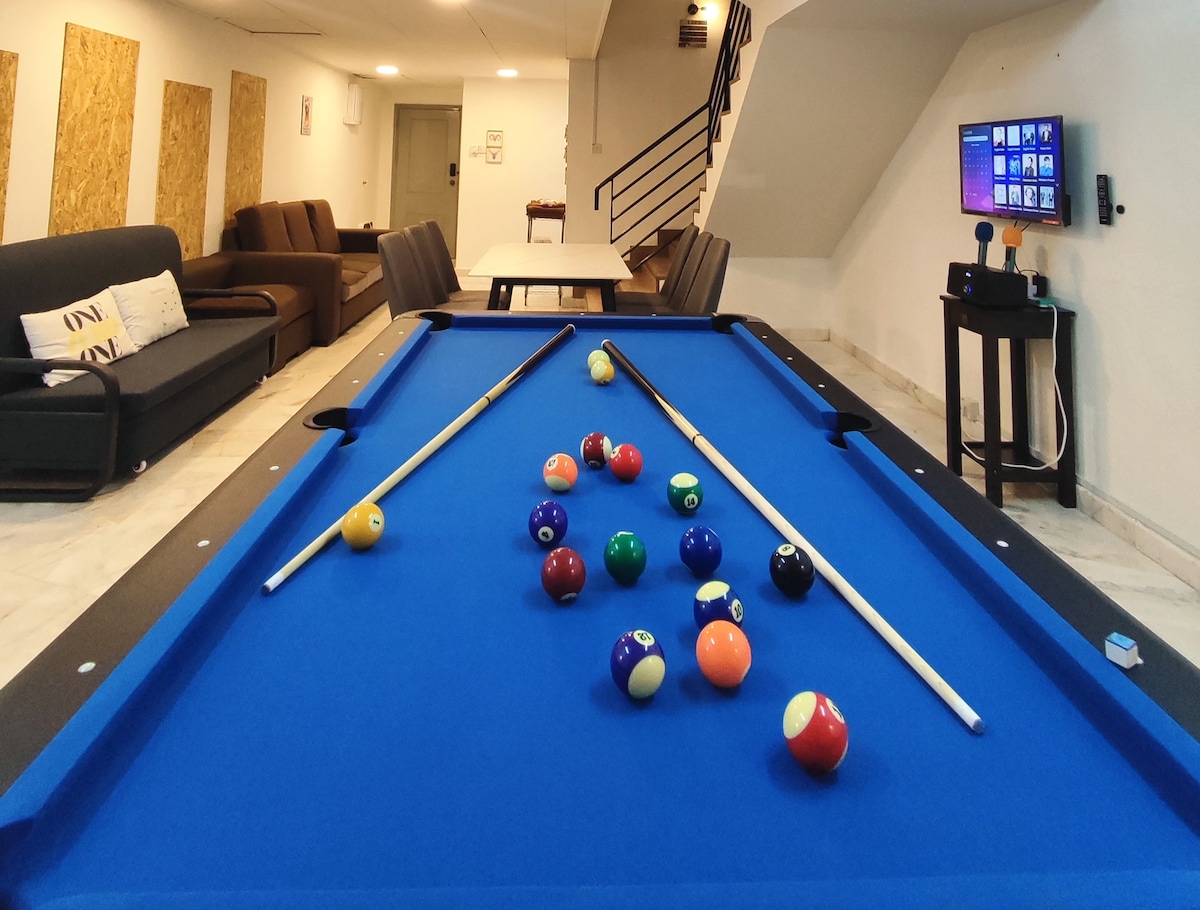 A vibrant game room is featured with a billiards table at its center, adorned with colorful pool balls. Surrounding seating areas include a dark sofa and a dining table. A flat-screen TV is mounted on the wall, creating a relaxed atmosphere for entertainment.
