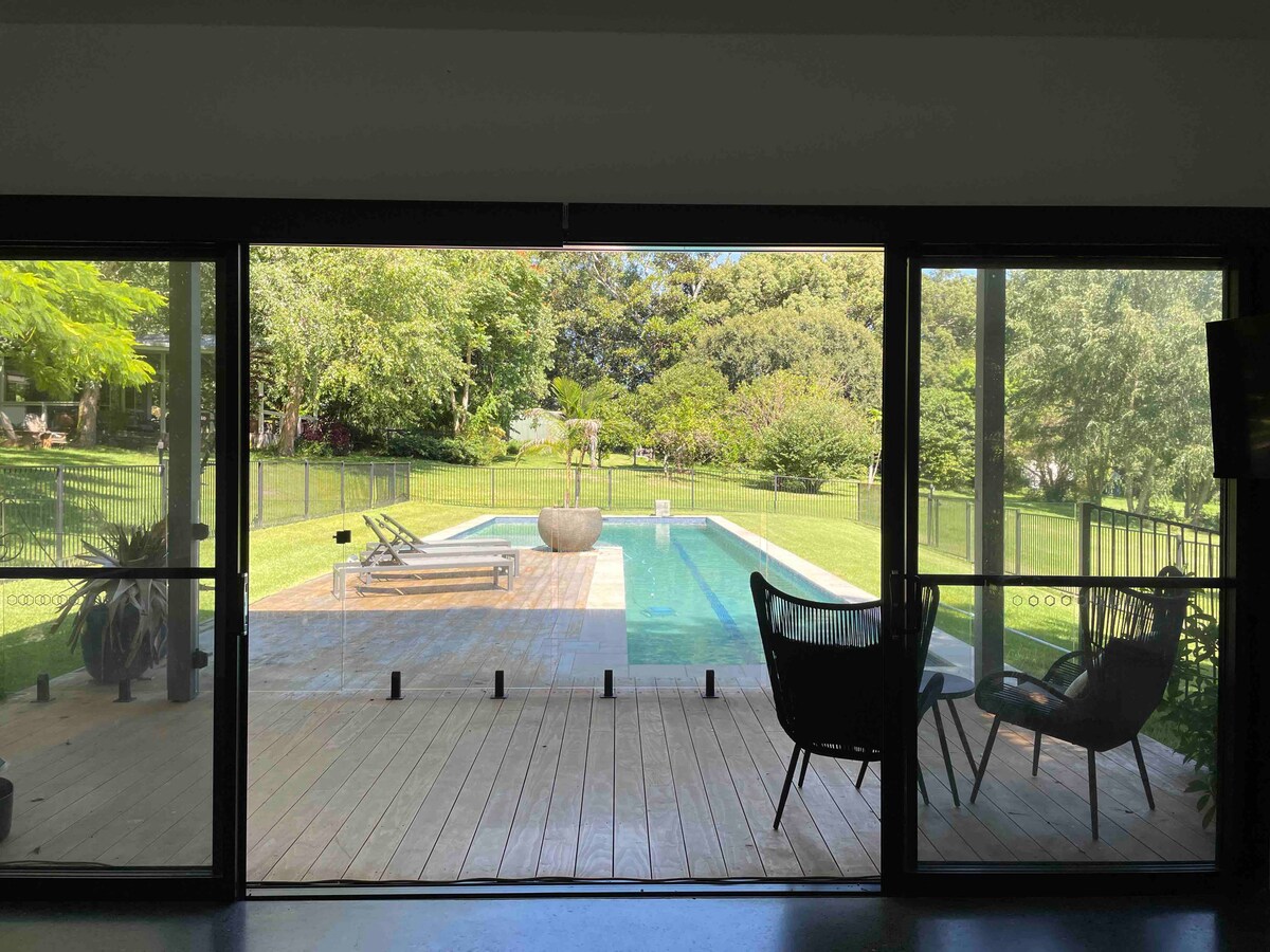 A view through large glass doors reveals a serene outdoor space featuring a heated lap pool framed by landscaped grass. Comfortable chairs are positioned on a wooden deck, providing a relaxing spot to enjoy the surroundings that include lush greenery and trees.