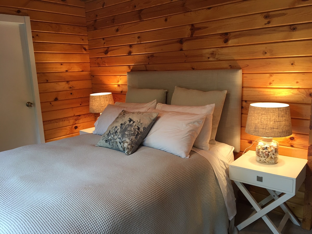 A cozy bedroom features a spacious bed dressed in light grey linens, complemented by wooden wall panels. Two bedside tables with soft lamp lighting create a warm ambiance. A decorative pillow adds a subtle touch to the bedding, enhancing the room's inviting feel.