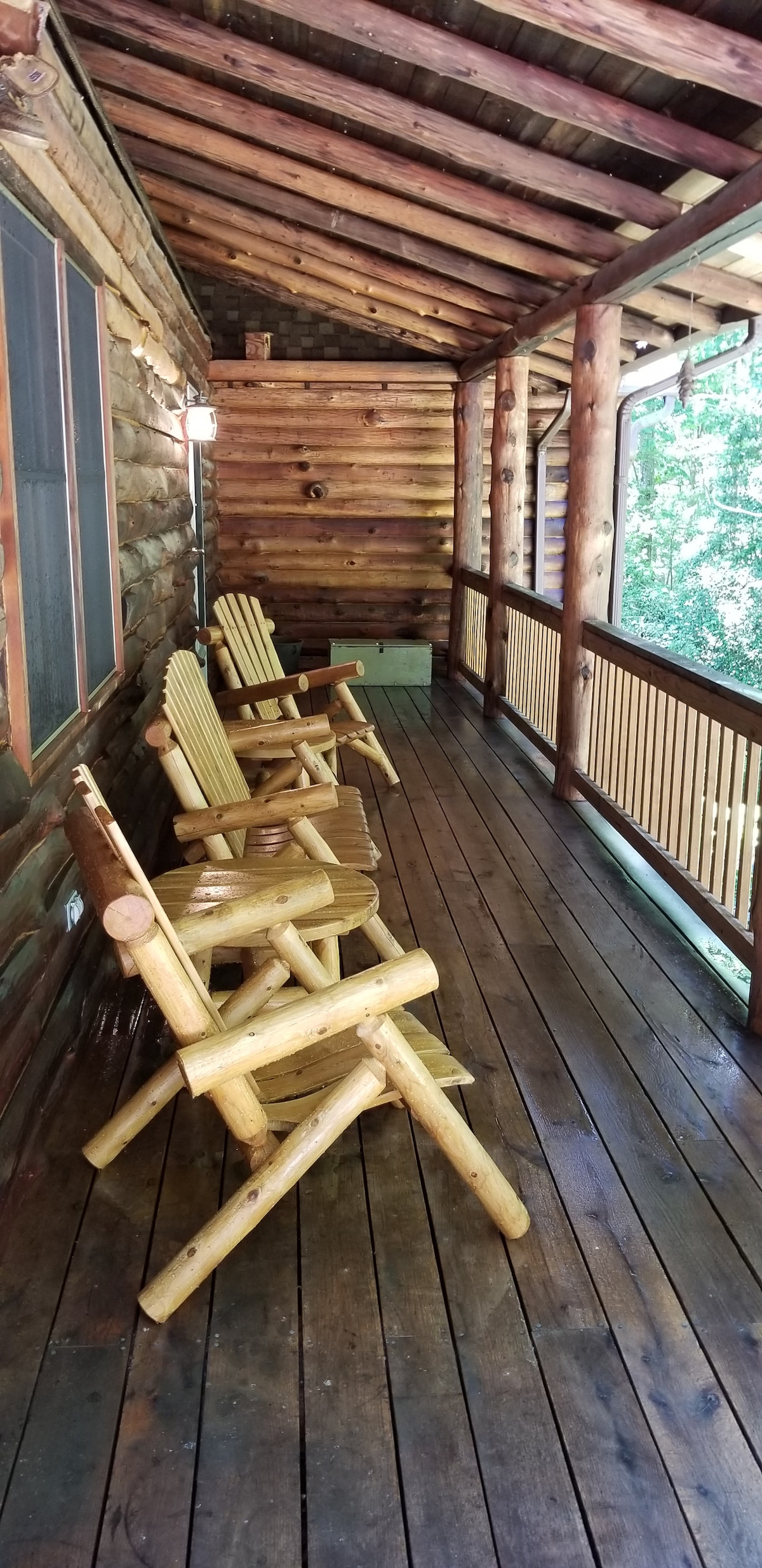 A covered porch features several rustic wooden rocking chairs positioned along the natural wood flooring. Logs from the cabin's structure are visible, contributing to the cabin's cozy feel. Surrounding trees provide a sense of privacy and connection to nature.