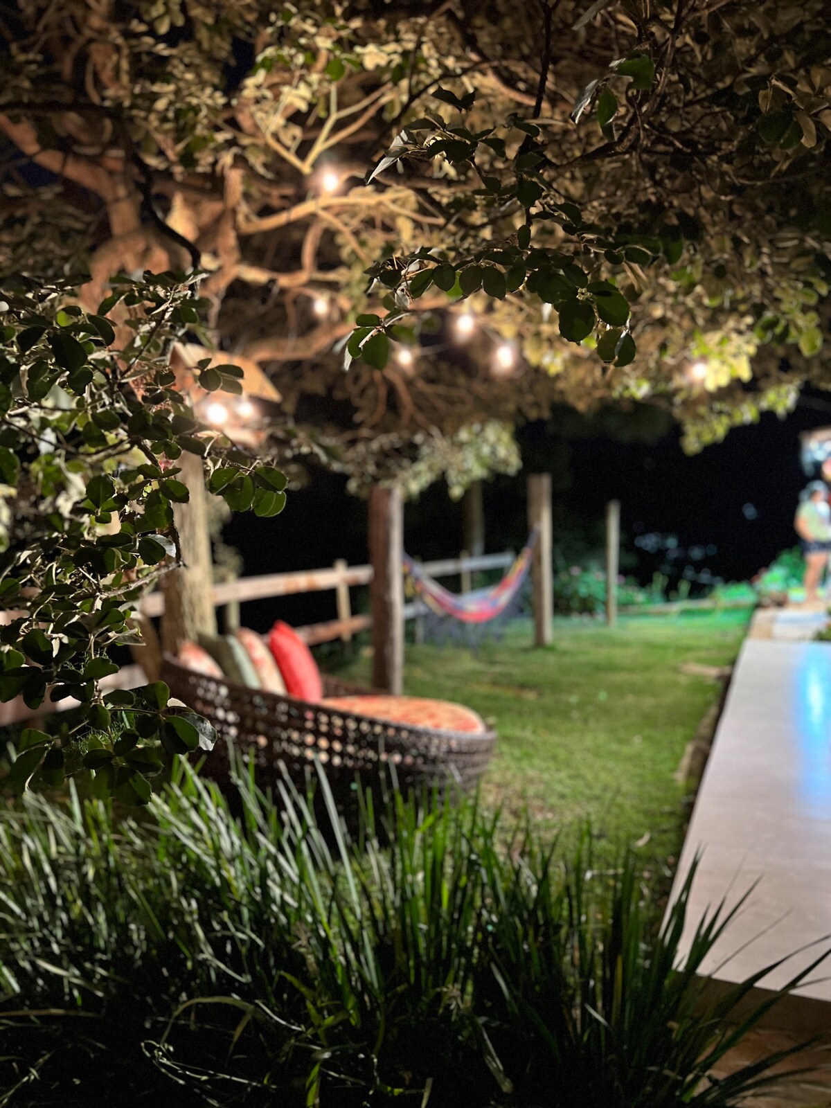 A serene outdoor area is illuminated by hanging lights, featuring a cozy seating arrangement with cushions. A hammock is gently swaying in the background, surrounded by lush greenery and a well-maintained lawn that leads to open space.