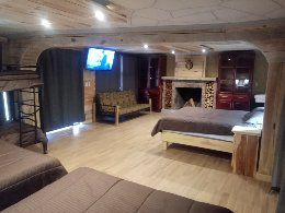 A spacious room features wooden paneling and soft lighting, creating a cozy atmosphere. A large bed is positioned next to a comfortable sofa. A TV is mounted on the wall, with a fireplace visible nearby. Bunk beds add additional sleeping options.