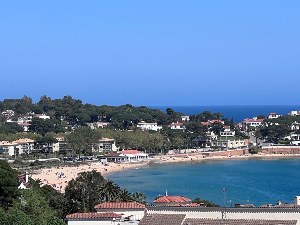A scenic view of Sant Pol beach is presented, showing golden sand populated with beachgoers. Lush green trees and residential buildings border the beach, framed by the shimmering blue sea under a clear sky. The colors create a soothing coastal atmosphere.